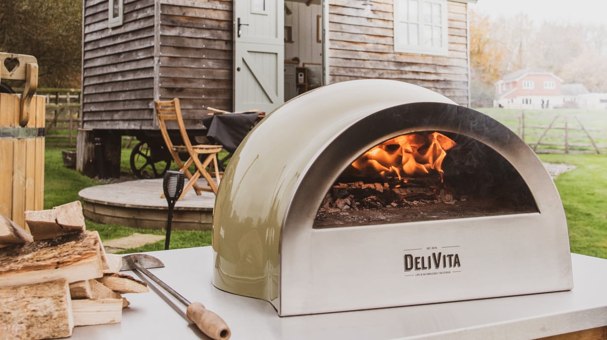 DeliVita pizza oven sat on a stainless steel outdoor table infant of a cabin and decking